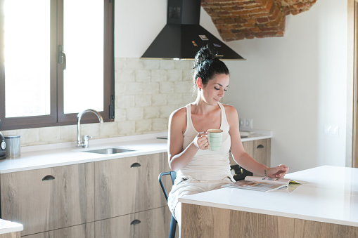 young brunette girl reading a magazine while drinking coffee in the kitchen