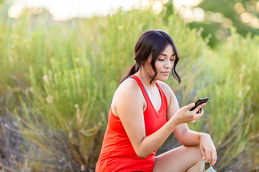 In Western Colorado One Attractive Generation Z Female of Hispanic Ethnicity Using Technology of smart phone Outdoors while crouching in front of a bush taking a rest from walking and jogging (Shot with Canon 5DS 50.6mp photos professionally retouched - Lightroom / Photoshop - original size 5792 x 8688 downsampled as needed for clarity and select focus used for dramatic effect)