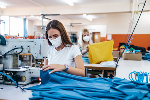 Professional seamstresses with face protective masks work in tailoring or sewing industry production hall.