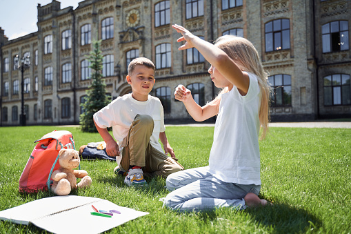 Cute boy and girl are sitting on grass with pencils and toys on sunny warm day