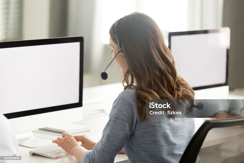 Call center operator using computer, looking at screen mock up Call center operator in wireless headset with microphone using computer, looking at blank white screen mock up, busy female employee working in customer support service office, consulting client Real Estate Agent Stock Photo