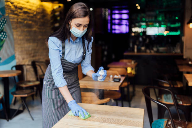 Young Caucasian female waitress cleaning the tables Young Caucasian female waitress wearing an apron, face mask and gloves, cleaning the tables using a wet wipe and a sanitizer restaurant cleaning stock pictures, royalty-free photos & images