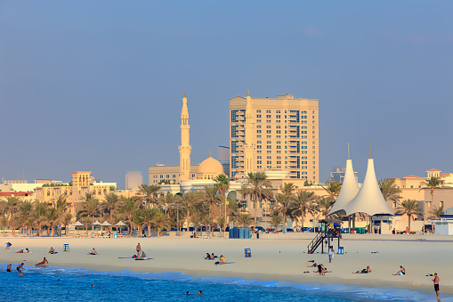 Dubai, United Arab Emirates - November 27, 2011: Tourists and local residents on Jumeirah Beach; some in the sea, swimming or wading. Others walk on the beach. In the background a dome and two minarets of the Jumeirah Grand Mosque can be seen. Photo shot in the late afternoon sunlight; copy space.