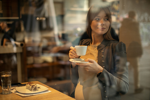 Beautiful young pregnant woman enjoying fresh coffee and chocolate cake in coffee shop while sitting next to the window and watching through. She is in middle trimester and looks very satisfied and happy.
