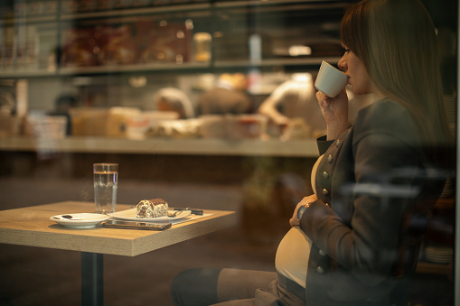 Beautiful young pregnant woman enjoying fresh coffee and chocolate cake in coffee shop while sitting next to the window. She is in middle trimester and looks very satisfied and happy.