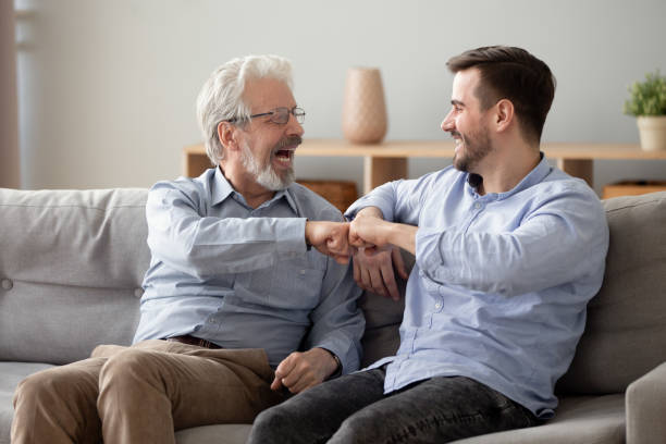 hombre de los años 60 y hombre milenario dando golpe de puño sentado en el interior - adult offspring family multi generation family senior adult fotografías e imágenes de stock