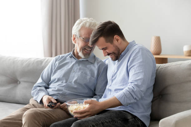 Elderly father grownup son laughing eating popcorn enjoy weekends together Elderly 60s father grown up son sitting on couch watching comedy movie laughing feels overjoyed eating pop corn snack enjoy weekend together at home, daddy and adult child connection and love concept divergent movie review stock pictures, royalty-free photos & images