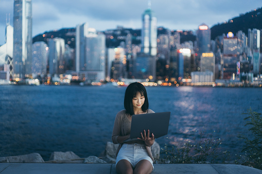 Young Asian woman using laptop by the promenade of Victoria harbour, against illuminated Hong Kong cityscape at dusk