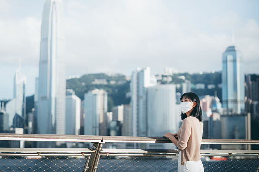 Young Asian woman wearing a protective face mask to protect and prevent from the spread of viruses in the city, looking away while standing by the promenade of Victoria harbour against Hong Kong city skyline