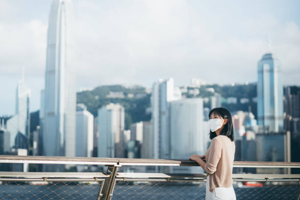 young asian woman wearing a protective face mask to protect and prevent from the spread of viruses in the city, looking away while standing by the promenade of victoria harbour against hong kong city skyline - china covid imagens e fotografias de stock