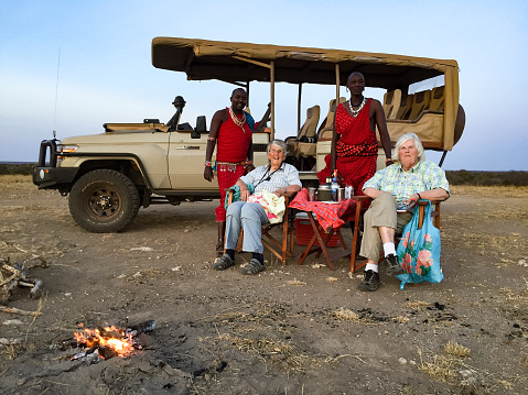 Senior female tourists with guides having a sundowner i Masai Mara. They are sitting in front of the landrover on the dry savannah.