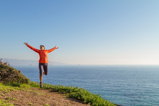 Joyful and Cheering latin mature woman enjoying the freedom with arms outstretched under sunrise at the beach, wearing a orange coat and short training pants. On background, Pacific Ocean landscape view.

Fitness. Healthy lifestyle.