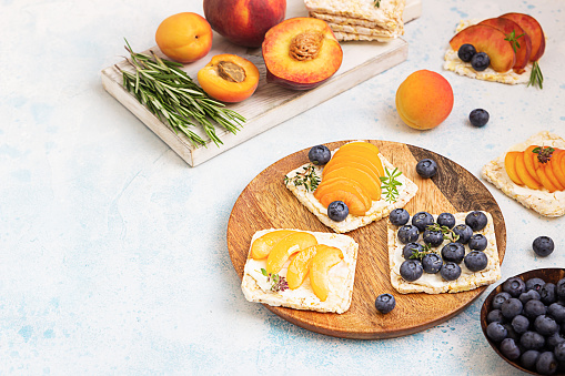 Mini rice cakes with blueberries, apricots, nectarines and cream cheese with honey for healthy breakfast. Light blue concrete background. Summer dieting sandwiches.