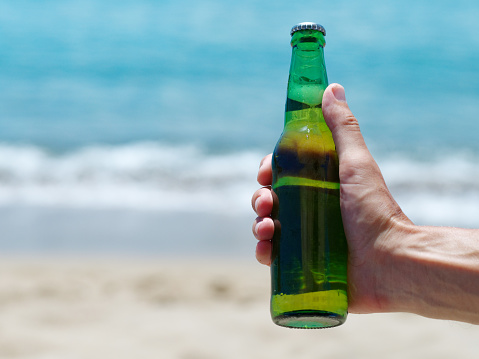 Male hand holding bottle of beer against ocean sandy beach. Copy space.