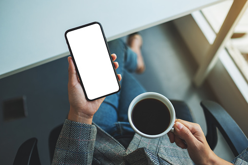 Top view mockup image of a woman holding mobile phone with blank white desktop screen with coffee cup