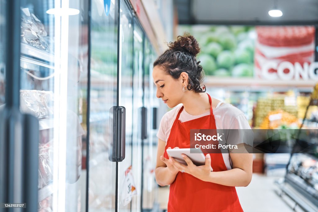 Saleswoman in apron working at supermarket Saleswoman working in supermarket. Female owner looking at refrigerator while holding digital tablet at store. She is wearing apron. Supermarket Stock Photo
