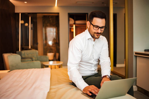 ritratto di un uomo affascinante che usa il laptop mentre è seduto sul letto nella camera d'albergo. - 4694 foto e immagini stock