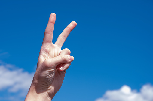Two fingers of a caucasian woman hand showing victory or peace gesture on blue sky background. Copy space.