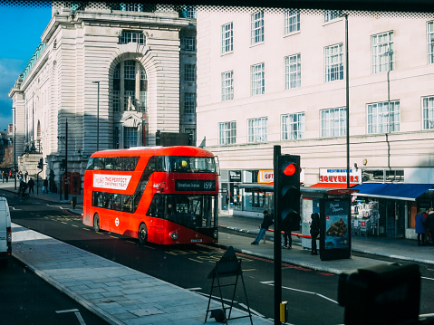 A double-decker bus drives down a street in London, England, with a historic building in the background. Tourists inspect the sights