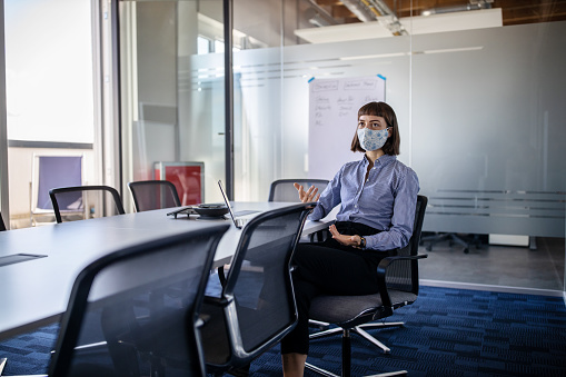 Businesswoman sitting at the office board room and talking on video call. Businesswoman with face mask video conferencing in office during pandemic.