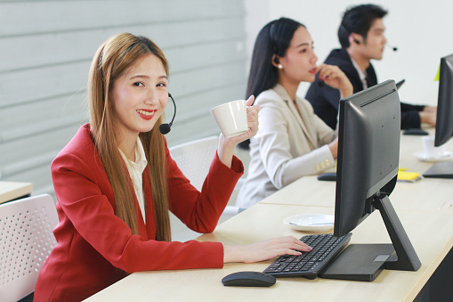 Smiling businesswoman Call Center hand holding a cup of hot coffee. Technical Support staff or receptionist phone operator teamwork. Asian customer support team.