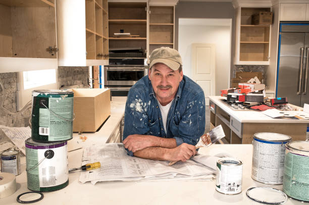 House painter in messy kitchen, with wet paint brush, looking at camera, in fixer upper house Male painter in clothes covered in paint and holding wet paint brush in messy home kitchen during remodel project with empty paint cans stacked everywhere on kitchen counter with lids off, kitchen cabinet doors open painting kitchen cabinets stock pictures, royalty-free photos & images