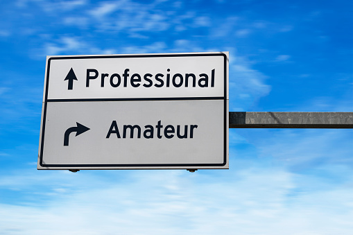 Professional versus amateur. White two street signs with arrow on metal pole. Directional road, Crossroads Road Sign, Two Arrow. Blue sky background.