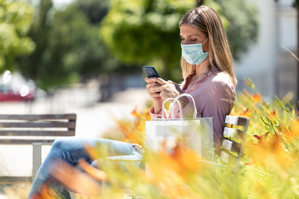 istock Beautiful blonde woman in face mask using her mobile phone while sitting on a bench in the street. 1263143076