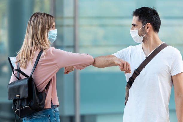 due amici che indossano la maschera chirurgica e sventolano con il gomito nel campus universitario durante l'epidemia di covid-19. distanziamento sociale e nuovo concetto normale. - men young adult human head blond hair foto e immagini stock