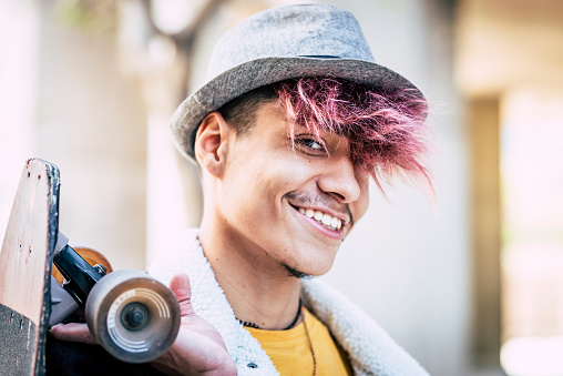 Portrait of handsome cheerful young man teenager boy smile and look at the camera - modern people with alternative look and colored hair - skateboard and hat on man