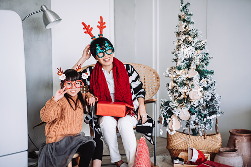 Joyful little Asian girl with grandmother wearing Santa Claus eyeglasses and reindeer headband having fun celebrating Christmas together in a Christmas party
