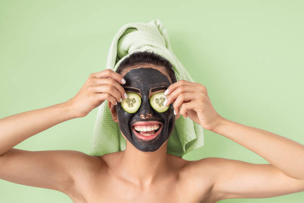 happy smiling girl applying facial carbon mask portrait - young woman having skin care cleanser spa day - healthy beauty clean treatment and and self care lifestyle concept - green background - facial mask spa treatment cucumber human face imagens e fotografias de stock