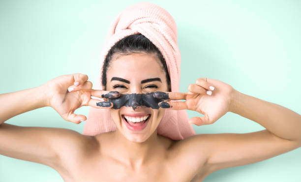 happy smiling girl applying facial charcoal mask portrait - young woman having skin care cleanser spa day - healthy beauty clean treatment and cosmetology products concept - aquamarine background - face mask imagens e fotografias de stock
