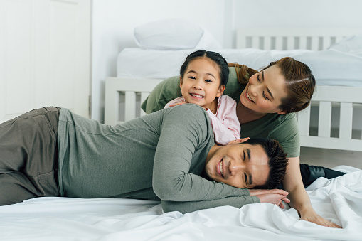 Cheerful Chinese mother and father with young daughter lying on bed