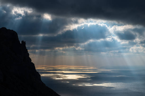 istock Sun rays break through dark clouds over the sea with a reflection and a black rock on the left 1263179537