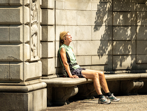 New York, NY, USA - August 1, 2020: A woman sits and rests in the sun with eyes closed at the East 90th Street and Fifth Avenue entrance (Engineers' Gate) to Manhattan's Central Park.