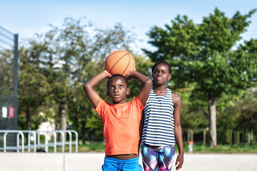 low angle view on african boy throwing basketball  for scoring on sports court at sunny weekend afternoon, his sister stands behind him