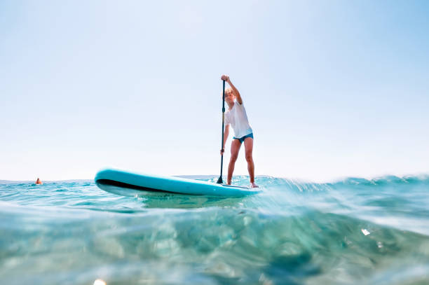 gülümseyen sarışın genç çocuk kürek tahtası ayağa su görüş açısı altında kürek kurulu. deniz konsepti görüntü yakınında aktif aile yaz tatili zamanı. - paddle surfing stok fotoğraflar ve resimler