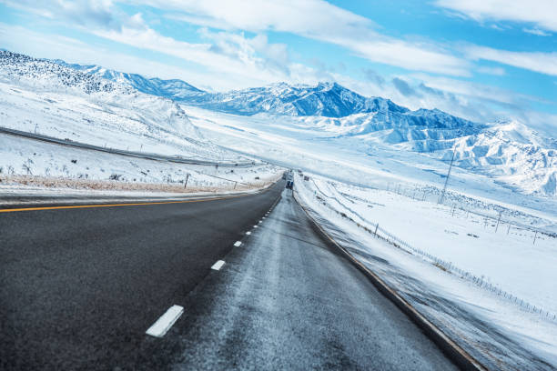 Long, diminishing perspective downhill southbound Interstate Route 15 expressway straightaway near Yuba State Park in southern Utah, western USA. Early February crosswinds blow snow sideways, creating a frozen and slippery asphalt road surface. The highway eventually disappears under a distant overpass, melting into the winter snow-blanketed valley plain beyond; with gradually rising foothills; then ultimately very high Wasatch Mountain Range peaks including Mount Nebo - the highest and southernmost Wasatch mountain peak in the far background. \