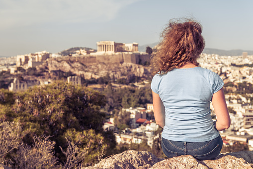 Young woman on background of urban landscape of Athens, Greece, Europe. Adult pretty girl tourist relaxes on hilltop overlooking Acropolis of Athens in summer. Concept of travel in Athens city.