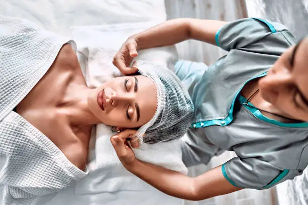 Young cute girl lying on a medical couch and getting ready putting on a hat. Beautician prepares the patient for a cosmetic procedure. Top view