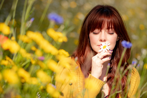 A beautiful red haired woman in a yellow cardigan sits in a field of yellow wild flowers. She holds a white flower against her face with her eyes closed as the sun shines in her face.