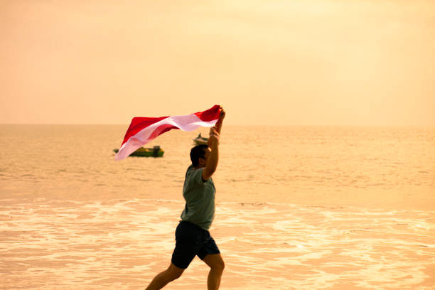 African woman in yellow outfit and headphones waving colorful scarf on beach.