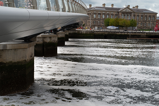 Belfast, Northern Ireland, UK - August 1, 2020:  Footbridge reaching over the River Lagan and its weir.  In the distance is the city's old Customs House.