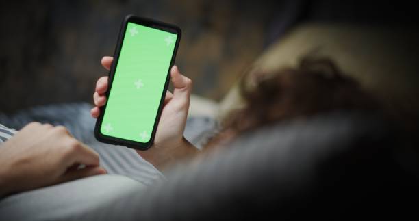 Woman is laying in bed and using smart phone. Close-up over shoulder view. Chroma screen on the phone la mujer está acostada en la cama y usando el teléfono inteligente. primer plano sobre la vista del hombro. pantalla de croma en el teléfono mujer está acostado en la cama y el uso de teléfono inteligente. pantalla de croma en el teléfono