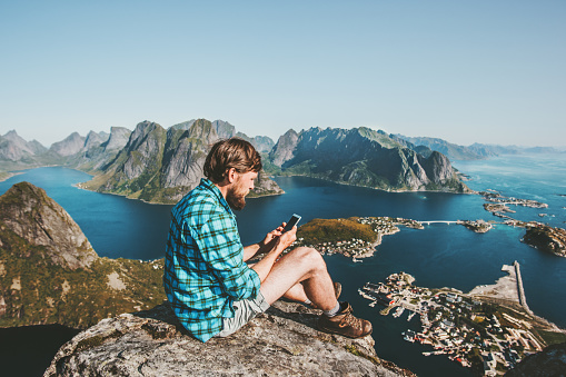 Man traveler using smartphone sitting on mountain top Traveling lifestyle concept adventure outdoor Reinebringen mountain aerial view Lofoten islands summer vacations in Norway