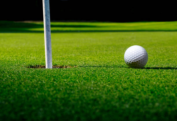 terrain de golf avec une balle à côté du trou et drapeau avec de longues ombres sur l’herbe verte. - sports flag flag shadow golf flag photos et images de collection