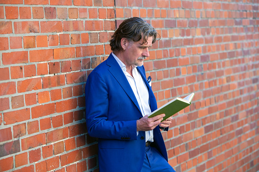 handsome businessman in his 50s standing at brick wall and reading a book
