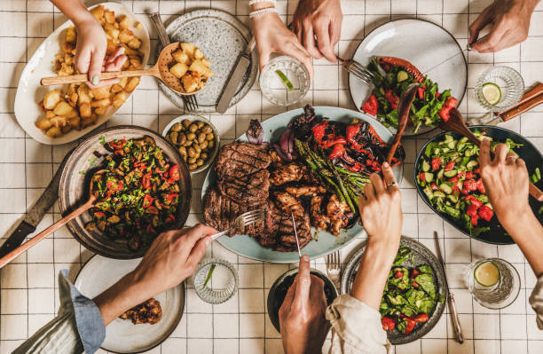 Flat-lay of people having barbeque party with meat and vegetables Summer barbeque party. Flat-lay of table with grilled meat, vegetables, salad, roasted potato and peoples hands with cutlery over white tablecloth, top view. Family gathering, comfort food concept people banque stock pictures, royalty-free photos & images
