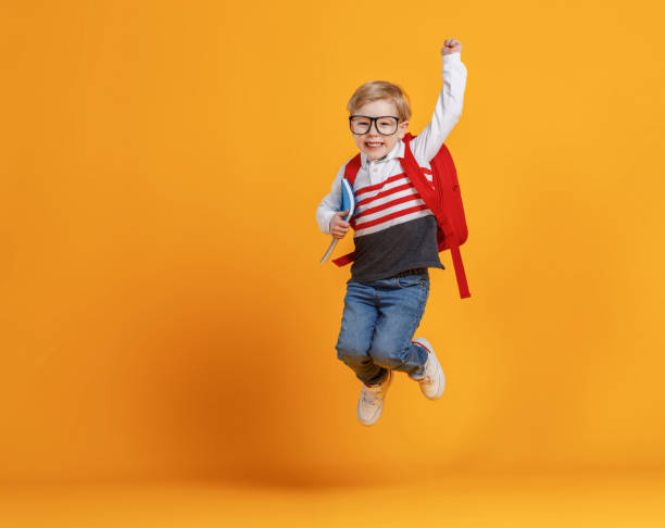 Happy school boy jumping with raised arm Full body delighted boy in trendy casual clothes smiling and jumping with raised arm while having fun before school studies against yellow backdrop schoolboy stock pictures, royalty-free photos & images